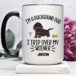 Pode incluir: Caneca de café de cerâmica branca com alça e borda pretas. A caneca apresenta um dachshund de desenho animado, uma tigela para cães, uma bola de tênis e o texto "Eu sou um pai de dachshund. Eu tropeço na minha salsicha. Jason".