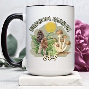Puede incluir: Taza de cerámica blanca con asa y borde negros. La taza presenta un gráfico de setas y helechos con el texto "Shroom Season 24-7".