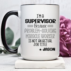 Puede incluir: Taza de café de cerámica blanca con asa y borde negros. La taza tiene texto negro que dice "I'm a Supervisor Because Problem-Solving Miracle Worker Is Not An Actual Job Title ★ JASON"