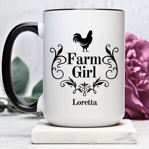 Könnte beinhalten: Eine weiße Keramiktasse mit schwarzem Henkel und Rand. Die Tasse zeigt eine schwarze Hahnensilhouette, den Text "Farm Girl" mit dekorativen Schnörkeln und den Namen "Loretta" aufgedruckt.