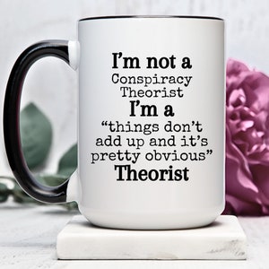 Puede incluir: Taza de café de cerámica blanca con asa negra. La taza tiene texto negro que dice "No soy un teórico de la conspiración Soy un "las cosas no cuadran y es bastante obvio" Teórico"