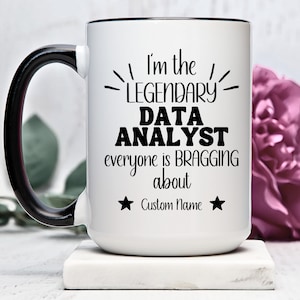 May include: White ceramic coffee mug with a black handle and black rim. The mug has a black graphic that says "I'm the Legendary Data Analyst everyone is bragging about" with a star on each side of the text and the words "Custom Name" in between the stars.