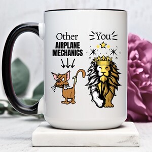 Pode incluir: Caneca de cerâmica branca com alça preta. A caneca tem uma borda preta e apresenta um gráfico de um leão usando uma coroa e um gato com um tapa-olho e uma bandagem na cauda. O texto na caneca diz "Other Airplane Mechanics" com setas apontando para baixo para o gato e "You" com estrelas acima do leão.