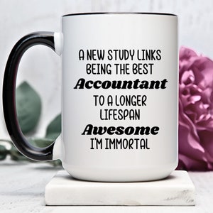 May include: White ceramic mug with a black handle and rim. The mug features the text "A NEW STUDY LINKS BEING THE BEST Accountant TO A LONGER LIFESPAN Awesome I'M IMMORTAL" in black lettering. A pink flower is in the background.