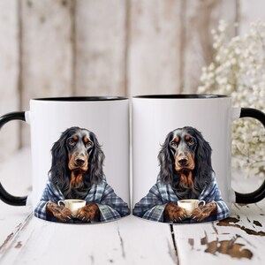 Könnte beinhalten: Zwei weiße Keramikbecher mit schwarzen Rändern und Griffen. Jeder Becher zeigt einen schwarzen und braunen Hund, der eine blaue karierte Decke trägt und eine kleine Tasse Kaffee hält.