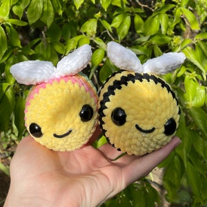 May include: Two crocheted bee plushies, one with rainbow stripes and white wings, the other with black stripes and white wings.