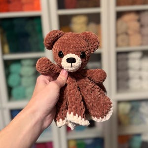 Puede incluir: Un oso de peluche de crochet marr&oacute;n con una manta de crochet blanca adjunta. El oso tiene ojos negros y una nariz negra.