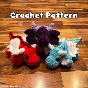 Puede incluir: Tres peluches de dragón de crochet en diferentes colores: rojo, morado y azul. Los dragones tienen detalles blancos y están tumbados sobre una superficie de madera. El texto "Crochet Pattern" está en la parte superior de la imagen.