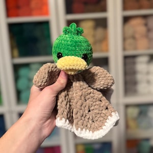 Puede incluir: Un pato de crochet verde con un pico amarillo y una manta de crochet marr&oacute;n unida. El pato est&aacute; siendo sostenido por una mano.