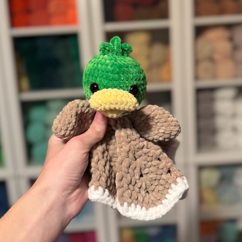 Puede incluir: Un pato de crochet verde con un pico amarillo y una manta de crochet marr&oacute;n unida. El pato est&aacute; siendo sostenido por una mano.