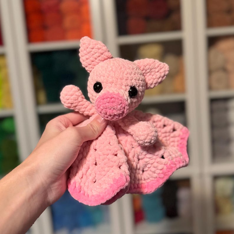 Puede incluir: Un cerdo de crochet rosa con una manta adjunta. El cerdo tiene ojos negros y un hocico rosa.