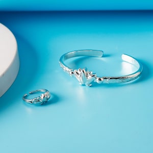 Könnte beinhalten: Ein silberfarbenes Armband- und Ring-Set. Das Armband ist im Manschettenstil gehalten und hat ein Blumendesign in der Mitte und geätzte Details. Der passende Ring hat ein ähnliches Blumendesign. Beide Teile sind auf blauem Untergrund.