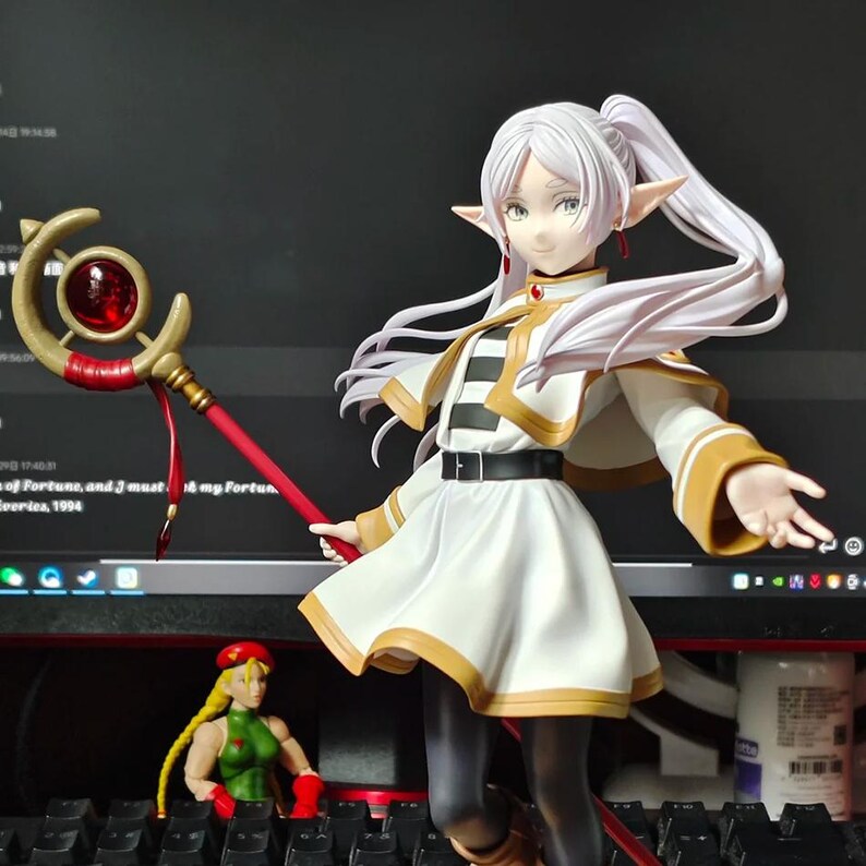 May include: A detailed figurine of a female anime character with long white hair in a ponytail, pointed ears, and a white and gold outfit. She holds a red staff with a large red gem. A smaller figurine of a female character in a green outfit is also visible.