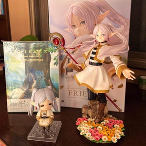 May include: A collection of anime-themed figurines and a soundtrack album. The main figurine is a character with long white hair, wearing a white and gold outfit, and holding a staff. A smaller figurine and album are also visible.