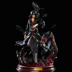 May include: A detailed figurine of a character in a long black coat with red accents and a red cloud symbol. The figure is surrounded by black birds and flames, set on a dark base. The character has black hair and is in a dynamic pose.