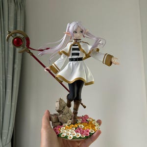 May include: A detailed figurine of a female anime character with long white hair and pointed ears. She wears a white dress with gold trim, black leggings, and brown boots. The figure holds a staff with a red gem and stands on a base with flowers.