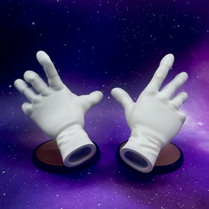 May include: Two white, open-palmed hands, each mounted on a small, round, brown base with a black rim. The hands are positioned against a purple and blue background with white speckles, resembling a starry sky.