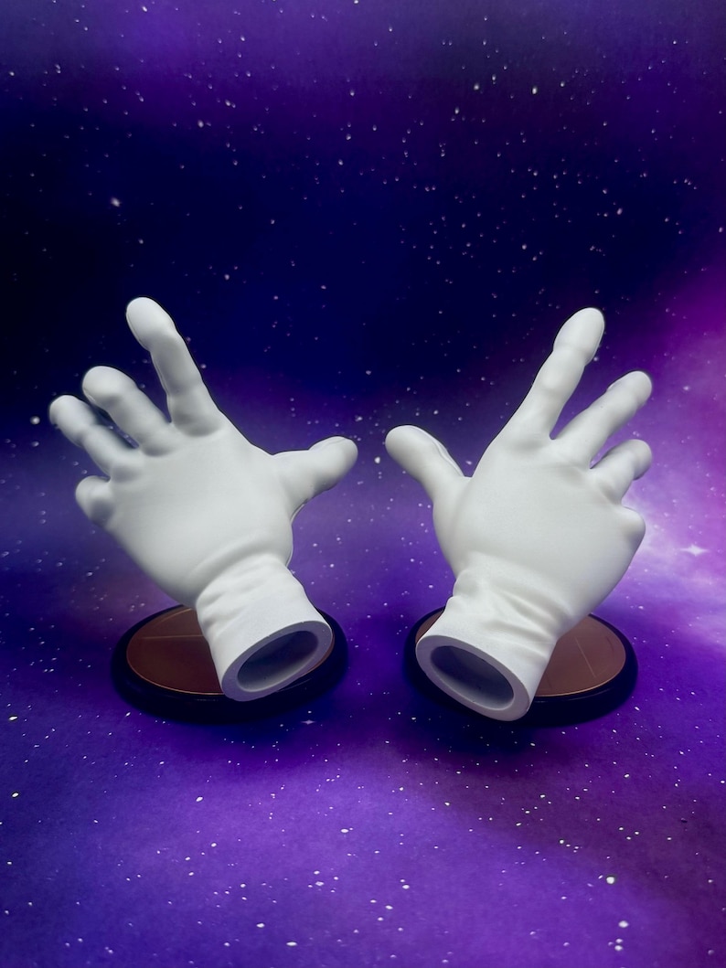 May include: Two white, open-palmed hands, each mounted on a small, round, brown base with a black rim. The hands are positioned against a purple and blue background with white speckles, resembling a starry sky.