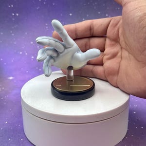 May include: A light blue hand figurine with a gold and black base, mounted on a white rotating platform. The hand is posed as if reaching out. The background is a purple gradient with white specks.