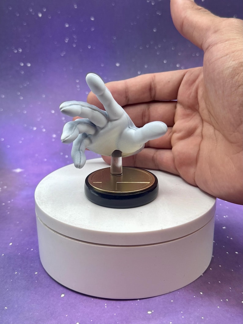 May include: A light blue hand figurine with a gold and black base, mounted on a white rotating platform. The hand is posed as if reaching out. The background is a purple gradient with white specks.