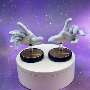 May include: Two light blue hand figurines with outstretched fingers, mounted on clear stands and black and gold circular bases. The hands are posed in a gesture. The figurines are displayed on a white platform against a purple background.