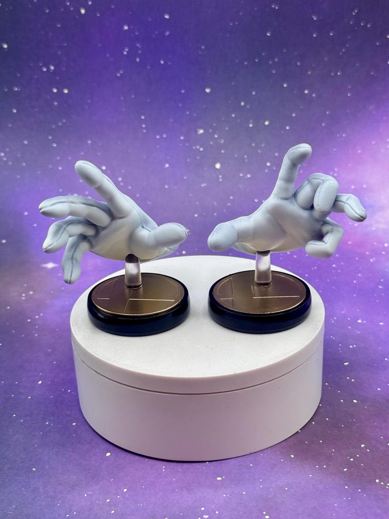 May include: Two light blue hand figurines with outstretched fingers, mounted on clear stands and black and gold circular bases. The hands are posed in a gesture. The figurines are displayed on a white platform against a purple background.