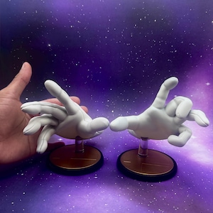May include: Two white hand sculptures, each mounted on a circular base with a gold-colored top and a black rim. The hands are posed in different gestures, set against a purple and blue starry background.