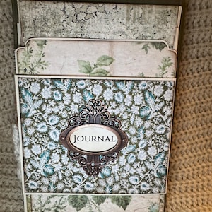 May include: A decorative journal with a floral pattern in shades of green and white. The journal features an ornate, oval-shaped label that reads "JOURNAL". The journal has a vintage aesthetic.