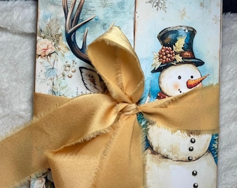 May include: A handmade journal with a winter theme. The cover features a watercolour illustration of a snowman and a deer with antlers. The journal is tied with a gold ribbon.