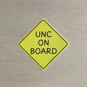 Puede incluir: Señal amarilla en forma de diamante con texto negro que dice "UNC ON BOARD". La señal tiene un borde negro y el texto "StickerRules" está impreso en letras pequeñas en la parte inferior.