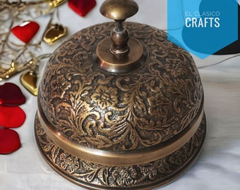 Op de afbeelding: Een vintage messingbel met een sierlijk bloemmotief. De bel heeft een afgeronde bovenkant en een brede basis. De bel staat op een wit oppervlak. De tekst "EL CLASICO CRAFTS" is zichtbaar op de achtergrond.