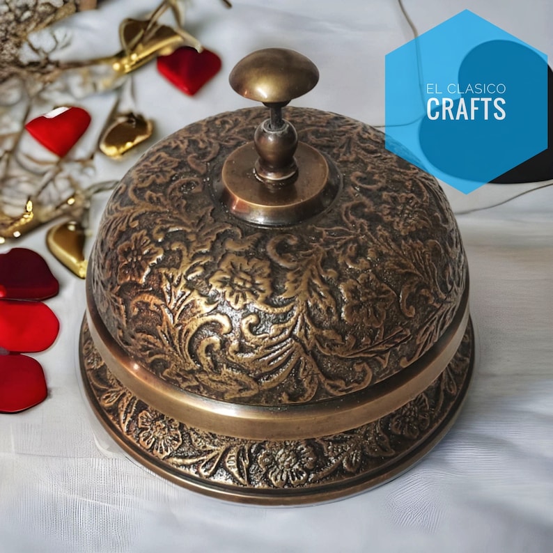 Puede incluir: Una campana de lat&oacute;n vintage con un dise&ntilde;o floral ornamentado. La campana tiene una parte superior redondeada y una base ancha. La campana est&aacute; sentada sobre una superficie blanca. El texto "EL CLASICO CRAFTS" es visible en el fondo.