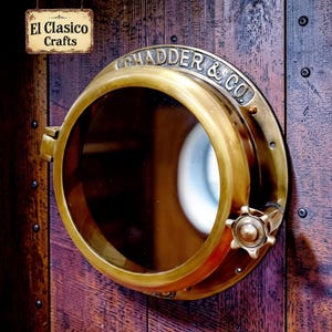 May include: A circular brass porthole window with a dark glass center, featuring the embossed text CHADDER & CO. along the top rim and a small star-shaped adjustment knob on the right side, mounted against a dark brown wooden plank wall.