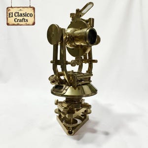 Op de afbeelding: Een landmeetkundig theodolietinstrument van gepolijst messing met een centrale telescoop, meerdere instelknoppen, een ronde basis en een driehoekige nivelleerplaat. Een bordje linksboven bevat de tekst El Clasico Crafts.