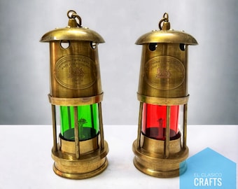 Op de afbeelding: Twee antieke messing mijnwerkerslampen met glazen schoorstenen. De lamp aan de linkerkant heeft een groene glazen schoorsteen en de lamp aan de rechterkant heeft een rode glazen schoorsteen. Beide lampen hebben een messing label met de tekst "Approved Flame Safety Lamp Made for Royal Navy London 1929".
