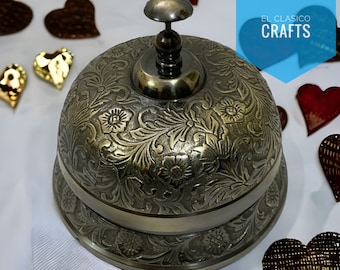 Puede incluir: Campana de latón antiguo con un diseño floral ornamentado. La campana tiene una parte superior redondeada con un pomo y una base ancha. La campana está rodeada de corazones decorativos.