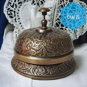 Puede incluir: Una campana de lat&oacute;n vintage con un dise&ntilde;o floral ornamentado. La campana tiene una parte superior redondeada y una base ancha. La campana est&aacute; sentada sobre una superficie blanca.