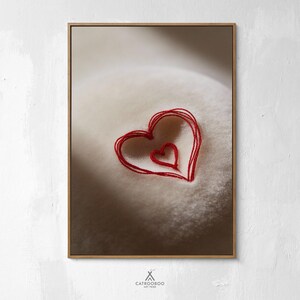 May include: A framed art print featuring a close-up of a textured, off-white surface with two red, hand-stitched hearts. The larger heart surrounds a smaller heart, creating a layered effect. The artwork is set against a soft, neutral background.