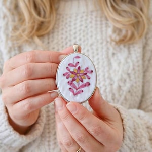 Op de afbeelding: Ovale hanger met een bloemenontwerp. De hanger heeft een witte achtergrond met een roze en geel bloemenpatroon. De hanger is omlijst in zilver. De hanger wordt voor een crèmekleurige trui gehouden.