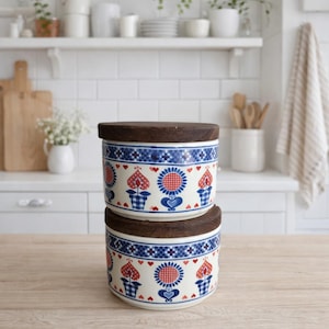 May include: Two ceramic canisters with wooden lids. The canisters are white with a blue and red floral pattern. The pattern includes hearts, flowers, and a checkered design. The canisters are stacked on top of each other.