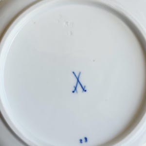 May include: The underside of a white porcelain plate featuring a blue underglaze crossed swords mark in the centre. The base also displays the blue painted number 23 and a faint, impressed numerical marking visible near the top edge of the rim.