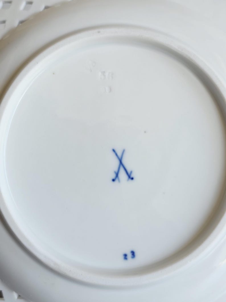 May include: The underside of a white porcelain plate featuring a blue underglaze crossed swords mark in the centre. The base also displays the blue painted number 23 and a faint, impressed numerical marking visible near the top edge of the rim.