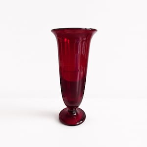 May include: A deep red glass vase featuring a flared, wide opening at the top that tapers down into a slender, cylindrical body. The vessel is supported by a circular, pedestal-style base, creating a smooth, translucent silhouette throughout.