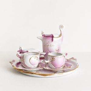 Puede incluir: Un juego de té de porcelana que incluye una tetera, dos tazas con platillos y un azucarero con tapa. Las piezas son blancas con degradados en rosa suave y detalles de volutas pintadas en oro, dispuestas sobre una bandeja redonda a juego.