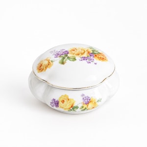 Könnte beinhalten: Eine runde Schmuckdose aus weißem Porzellan mit gewelltem Rand und abnehmbarem Deckel. Die Oberfläche ist mit einem Blumenmuster aus gelben Rosen und kleinen violetten Blüten verziert, das durch einen dünnen Goldrand am Rand akzentuiert wird.