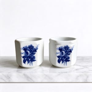 Könnte beinhalten: Zwei weiße Keramikbecher in sechseckiger Form, die jeweils auf der Vorderseite mit einem detaillierten blauen Blumenmuster aus Rosen und Blättern verziert sind. Die Becher haben eine glatte, glänzende Oberfläche und eine einfache Öffnung.
