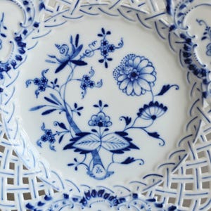 May include: A white porcelain plate featuring a blue onion pattern with floral motifs in the centre. The plate has a decorative lattice-style openwork border with blue painted accents along the edges and intricate floral designs throughout.