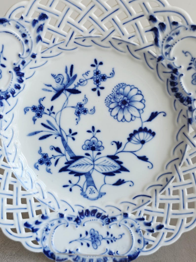 May include: A white porcelain plate featuring a blue onion pattern with floral motifs in the centre. The plate has a decorative lattice-style openwork border with blue painted accents along the edges and intricate floral designs throughout.