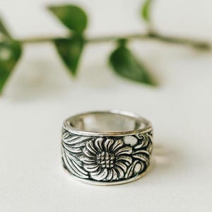 Puede incluir: Un anillo plateado con un diseño floral. El anillo presenta un girasol detallado en el centro, rodeado de hojas y patrones ondulados. El anillo es ancho y tiene un acabado ligeramente oxidado, lo que le da un aspecto vintage.