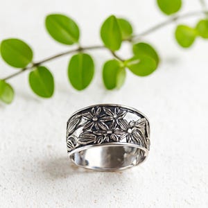 Puede incluir: Un anillo de plata con un diseño floral. El anillo presenta grabados detallados de flores y hojas alrededor de la banda. El anillo está sobre un fondo blanco con hojas verdes en el fondo.